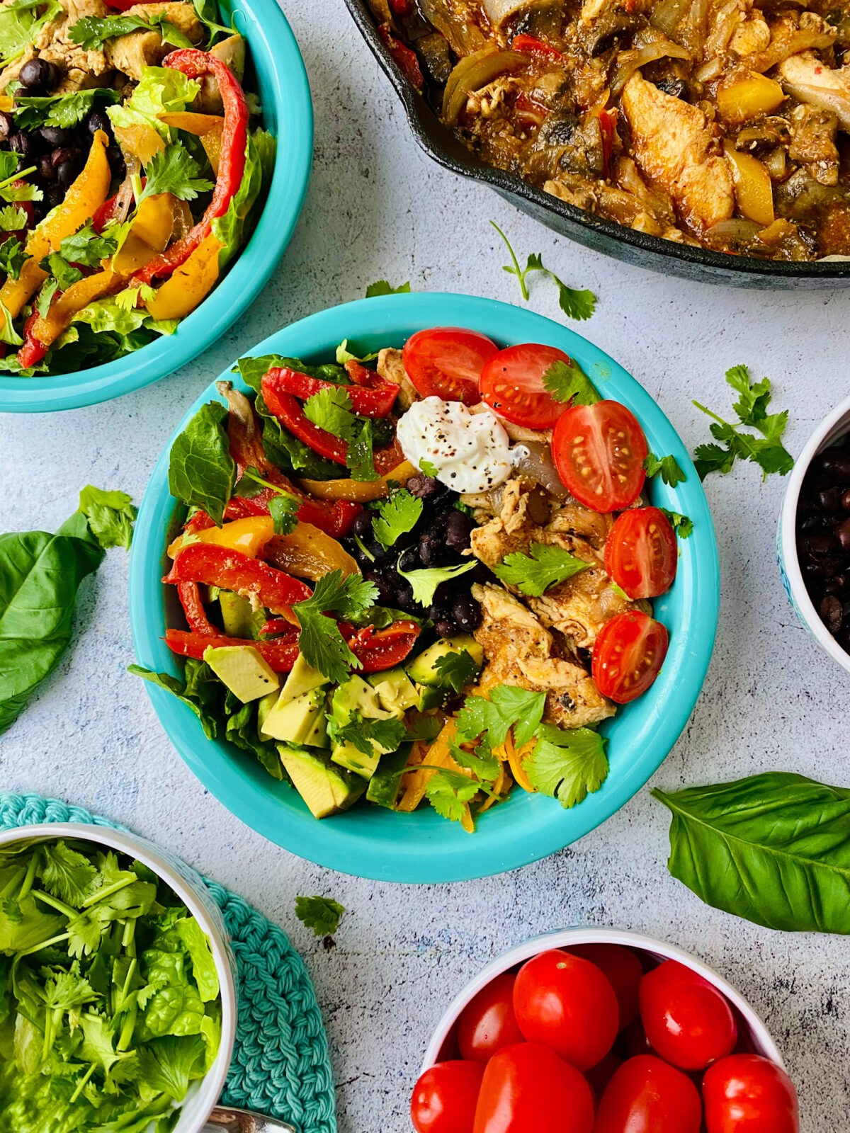 A picture of chicken fajita power bowl, prepared in their bowls with a cast iron pan full of fajita chicken. Other bowls surrounding the prepared dishes with extra ingredients surrounding the food subject as accessories to the picture.